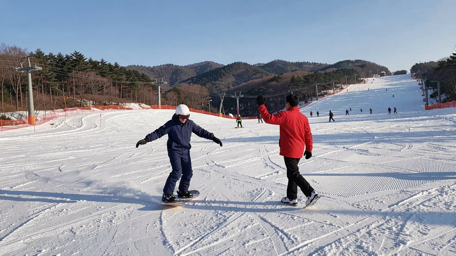 AI 생성 이미지: A first-time snowboarder in navy outerwear and a white helmet balances through a slow turn on a wide, gentle groomed slope while an instructor in a red jacket walks alongside with one hand raised, pine-covered ridges and a chairlift behind